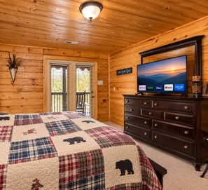 A cozy room with a wooden ceiling, a bed with a quilt, a dresser with a flat-screen TV displaying streaming services, and a window with blinds. - Mountain Views - 4 BR/4BA - 3 Mi to downtown Gatlinburg & Rocky Top Sports World (Gatlinburg)
