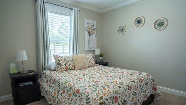 Quarto com colcha florida, mesa de cabeceira com abajur, criadeira com planta e janela com cortinas.