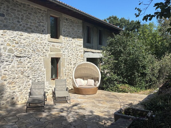 A stone building with a stone patio, two chairs, a large round wicker chair, and a tree.