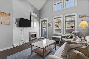 A living room with a fireplace, a flat-screen TV, and a sofa. - 2 bdrm penthouse with 180 Mountain view, heated pool & hot tubs (Canmore)