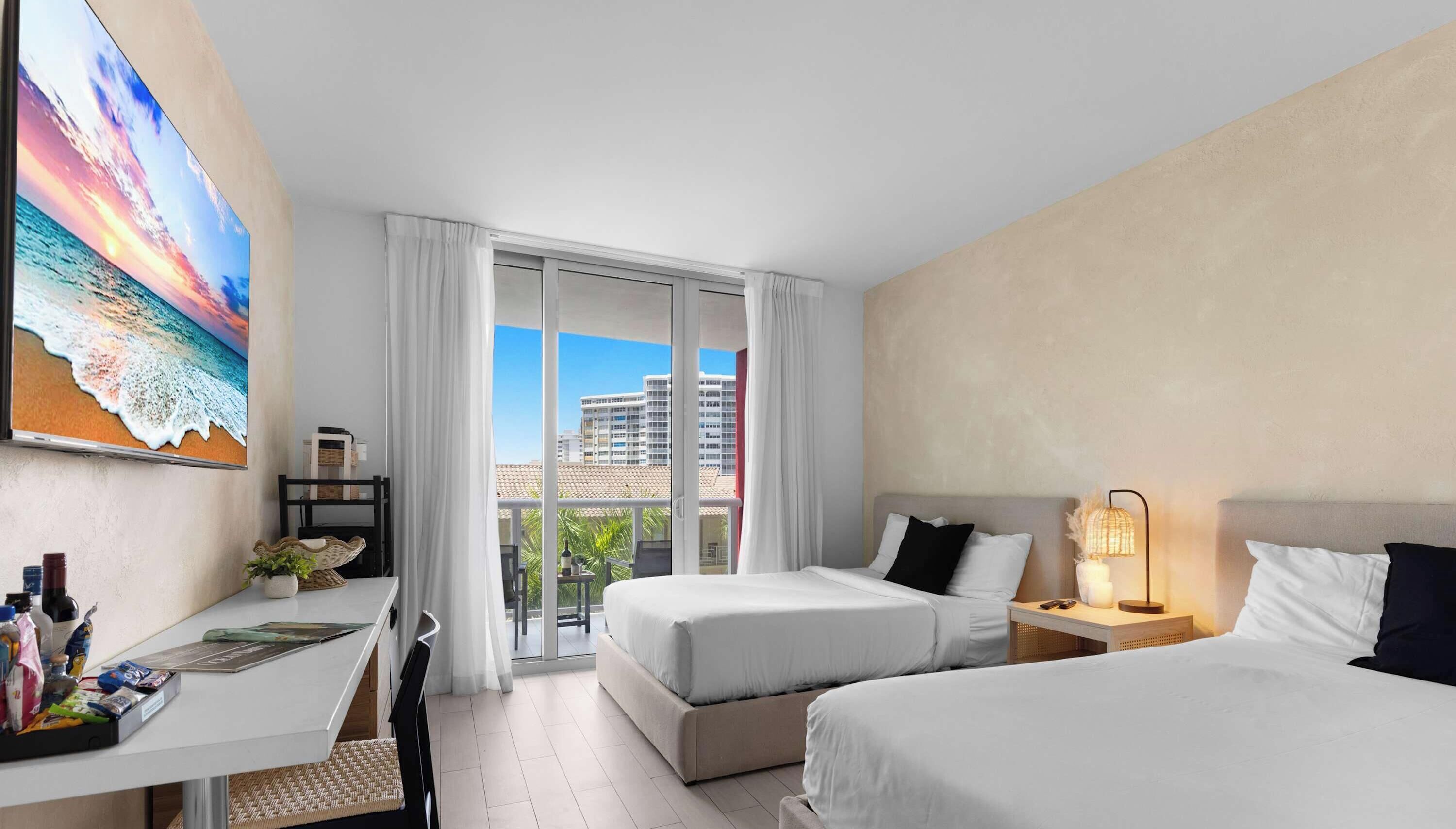 Ein Hotelzimmer mit zwei Betten, einem Schreibtisch, einem Stuhl, einem Balkon mit Blick auf Gebäude und einem Bild an der Wand.