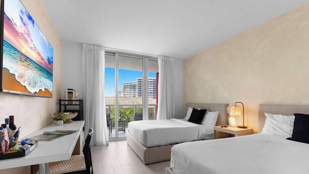Une chambre d’hôtel avec deux lits, un bureau, une chaise, un balcon donnant sur des immeubles et un tableau au mur.