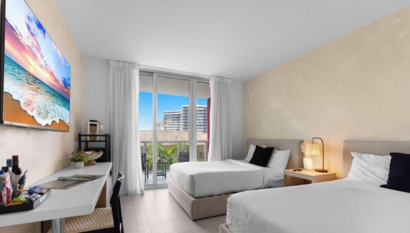 A hotel room with two beds, a desk, a chair, a balcony with a view of buildings, and a painting on the wall. - Beachwalk Cozy Studio w/ Balcony & Fitness Center (Hallandale Beach)