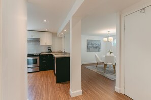 A modern kitchen with stainless steel appliances, a dining area with a white table and chairs, and a hallway with wooden flooring.