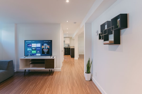 A modern living room with a flat-screen TV displaying a streaming service interface, a wooden floor, a white sofa, and a minimalist shelving unit.