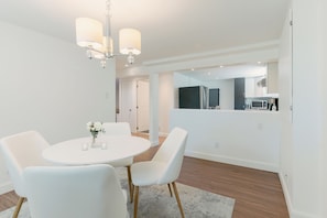 A modern dining area with a round table and white chairs, a chandelier, and a view into the kitchen.