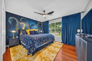 Ein Schlafzimmer mit Bett, Deckenventilator, Nachttisch, Kommode und einem Fenster mit Vorhängen.
