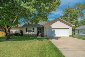 A single-story house with a front garage, a well-maintained lawn, and a large tree providing shade. - Lakefront Oasis: Pool, Fire Pit, Kayak, Paddle Boards, Game Room (Hot Springs)
