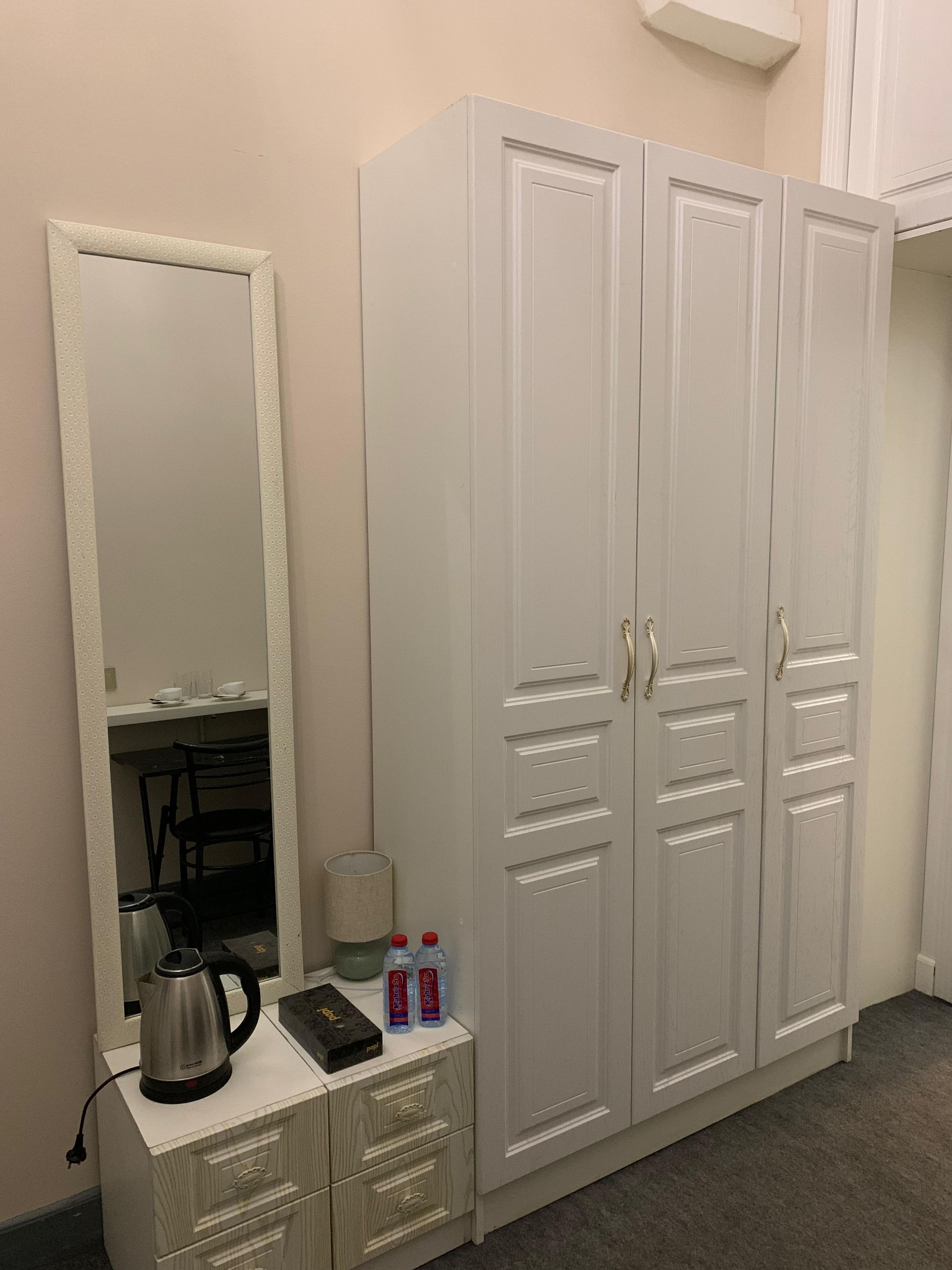 A room with a white wardrobe, a mirror, a desk with a kettle, and two bottles of water.