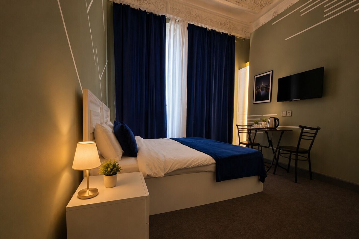 Una habitació d'hotel amb un llit, una taula de nit amb una làmpada, una petita planta, un escriptori amb una cadira i un televisor fixat a la paret.