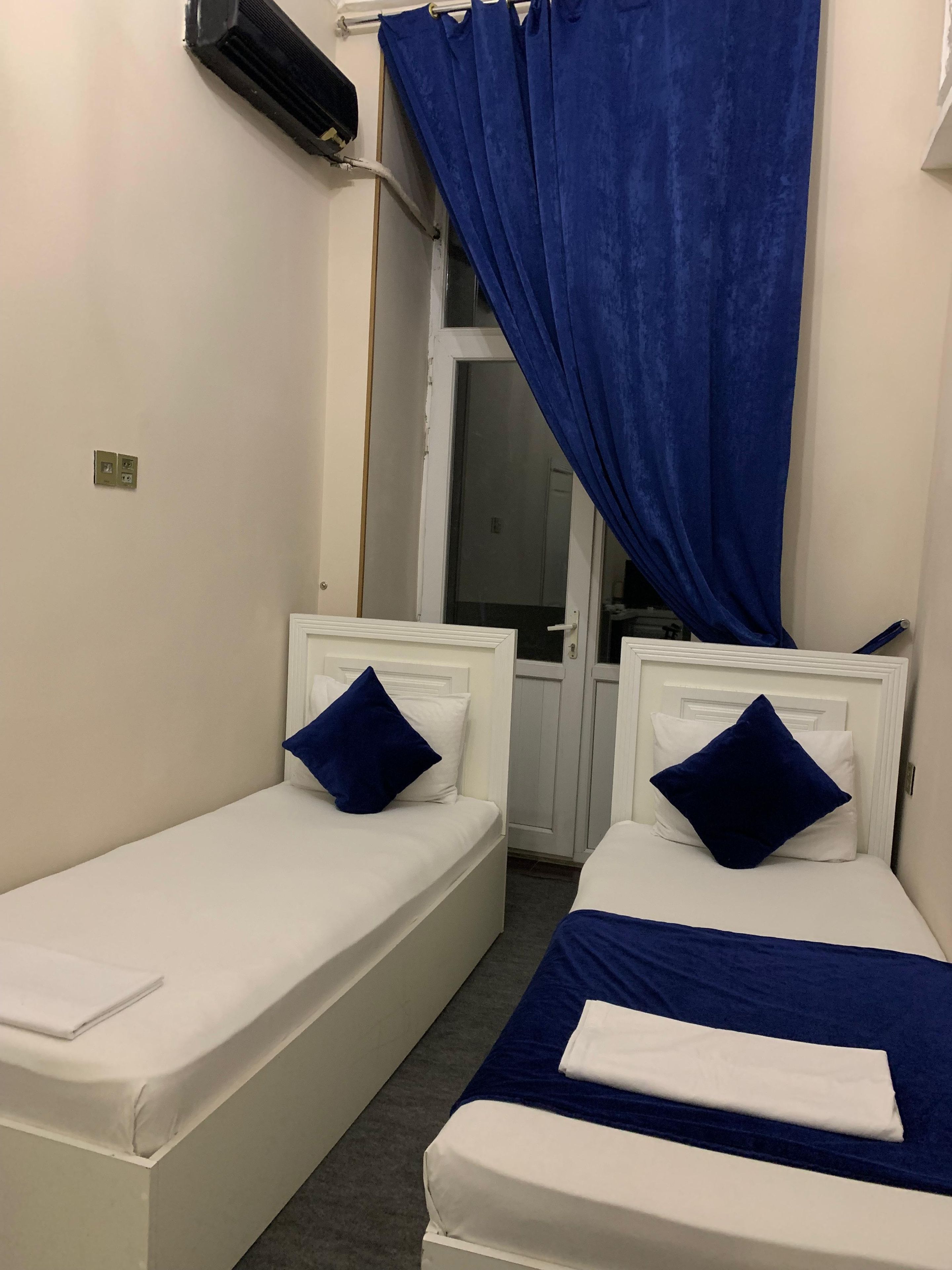 Una habitación pequeña con dos camas individuales, cada una con cabecero blanco y almohada azul. Se ve una ventana con cortinas azules y un equipo de aire acondicionado en la pared.