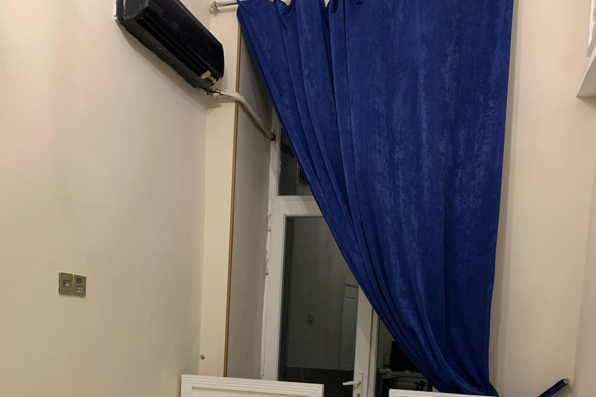 Una habitación pequeña con dos camas individuales, cada una con cabecero blanco y almohada azul. Se ve una ventana con cortinas azules y un equipo de aire acondicionado en la pared.