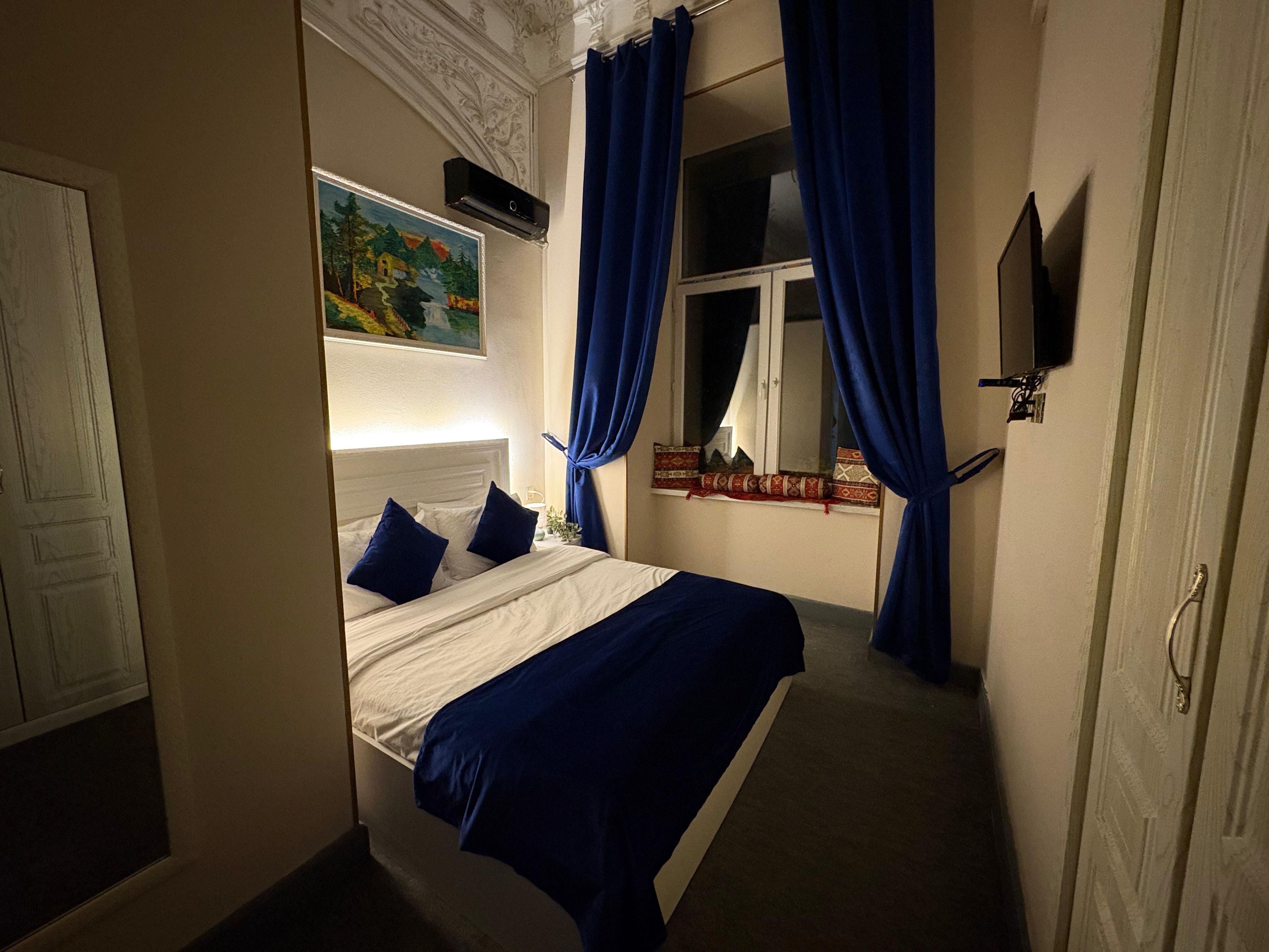 A bedroom with a bed, a painting, a window with curtains, and a wall-mounted TV.