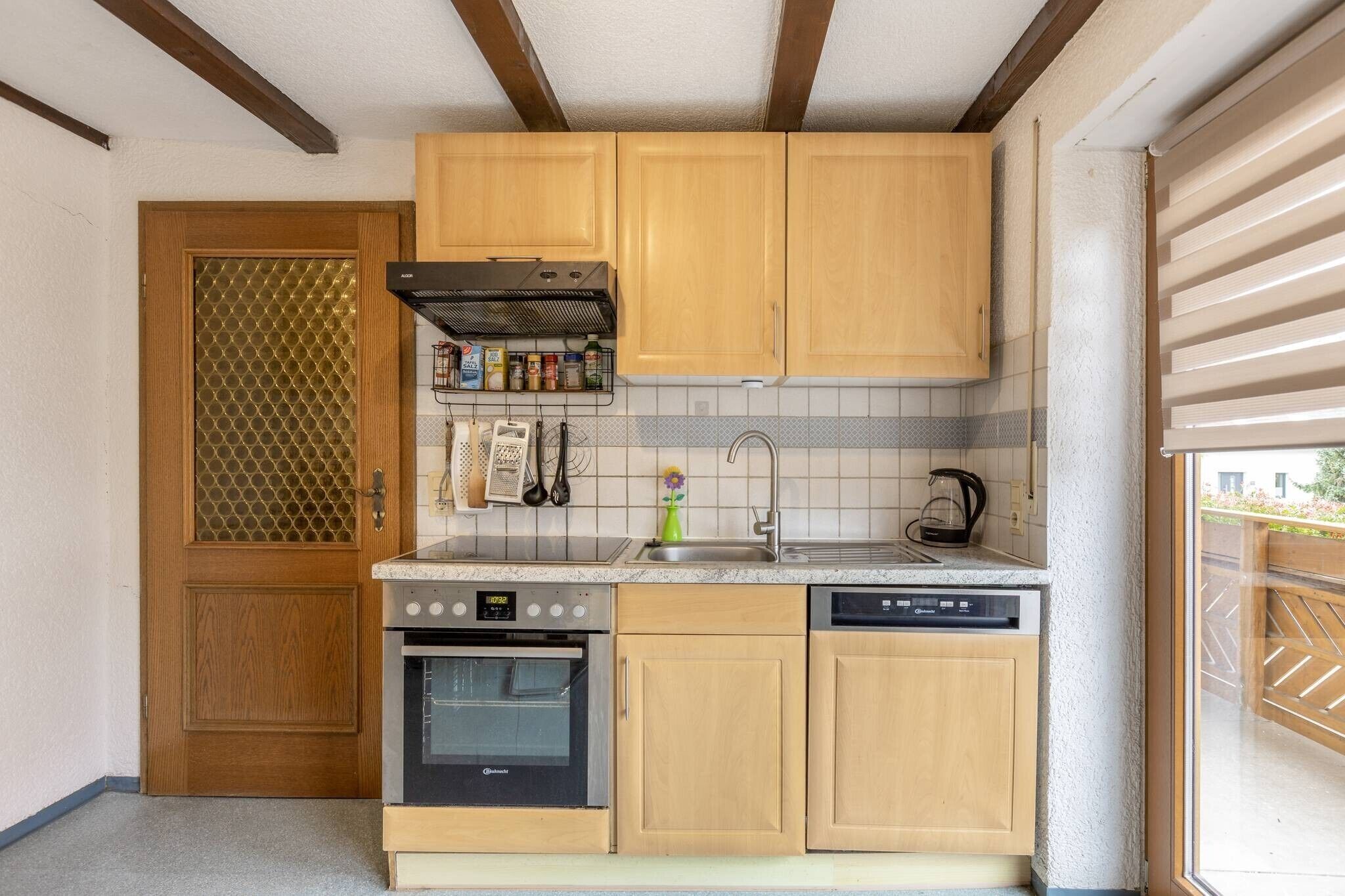 A compact kitchen with wooden cabinets, a built-in oven, a dishwasher, and a window with a view of the outside.