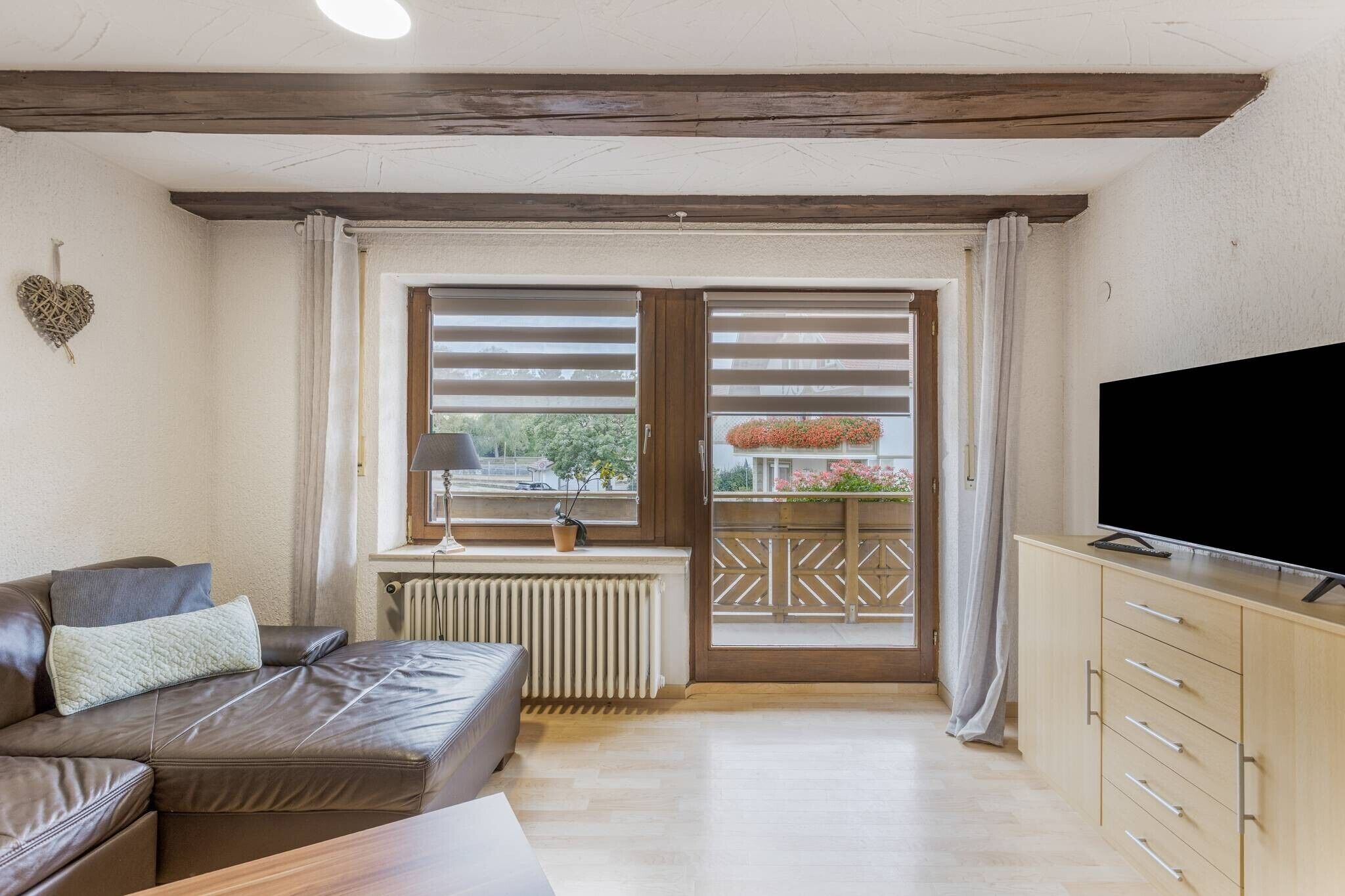 Ein Wohnzimmer mit Ledersofa, Fernseher, Heizkörper und einem Fenster mit Jalousien.