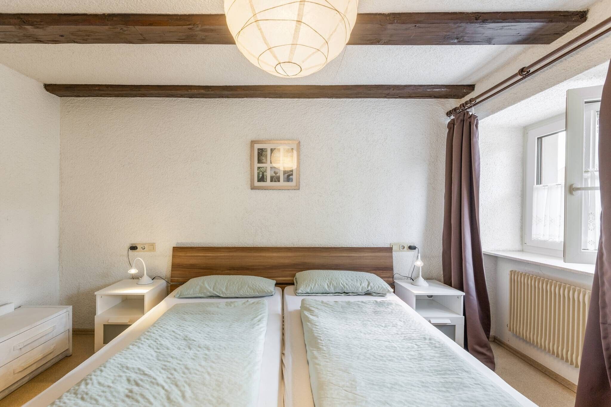 A room with two beds, a wooden ceiling, a wall-mounted picture, and a window with curtains.