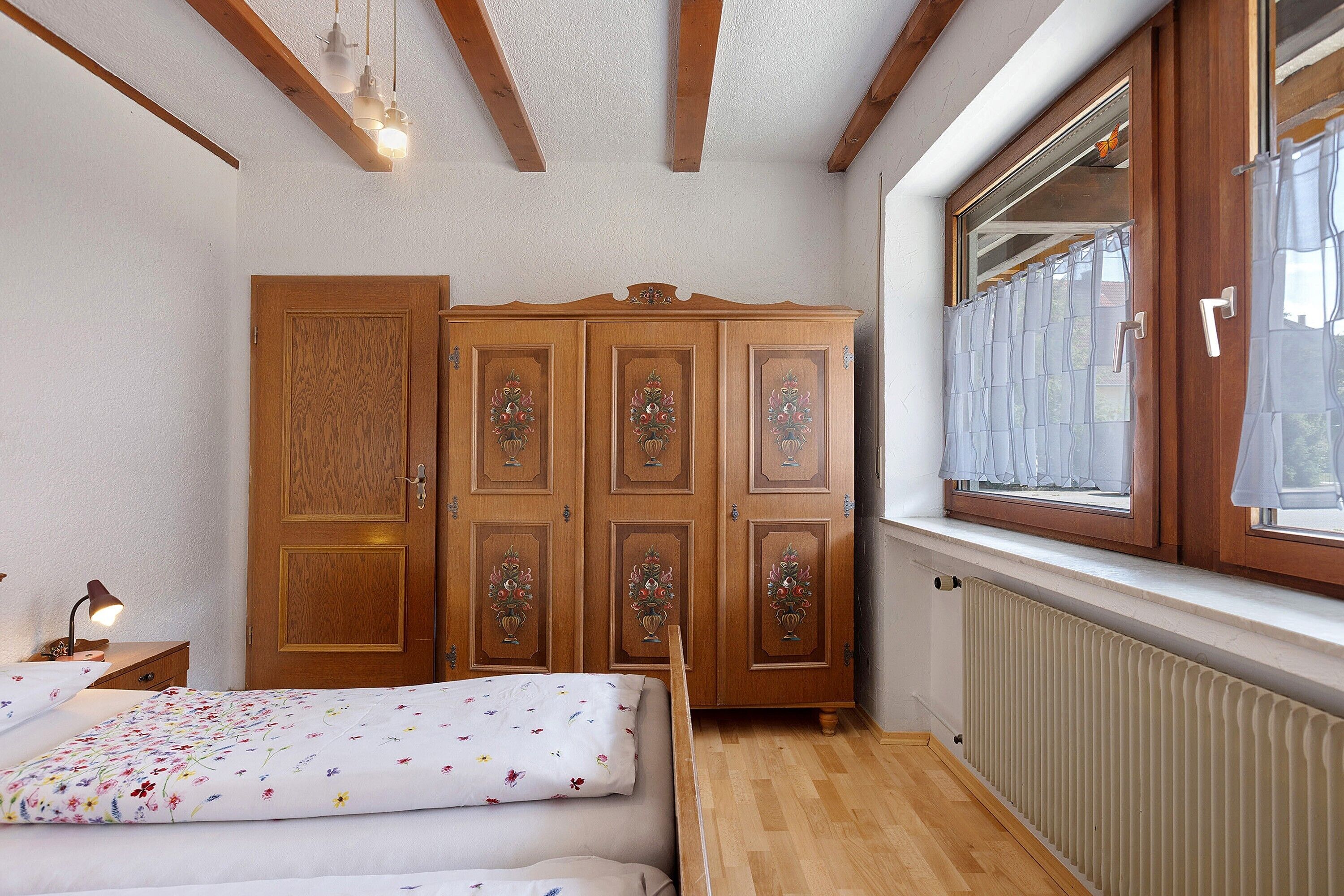 Ein Schlafzimmer mit Holzschrank, einem Bett mit floralem Bettbezug, einem Nachttisch und einem Fenster mit Blick.