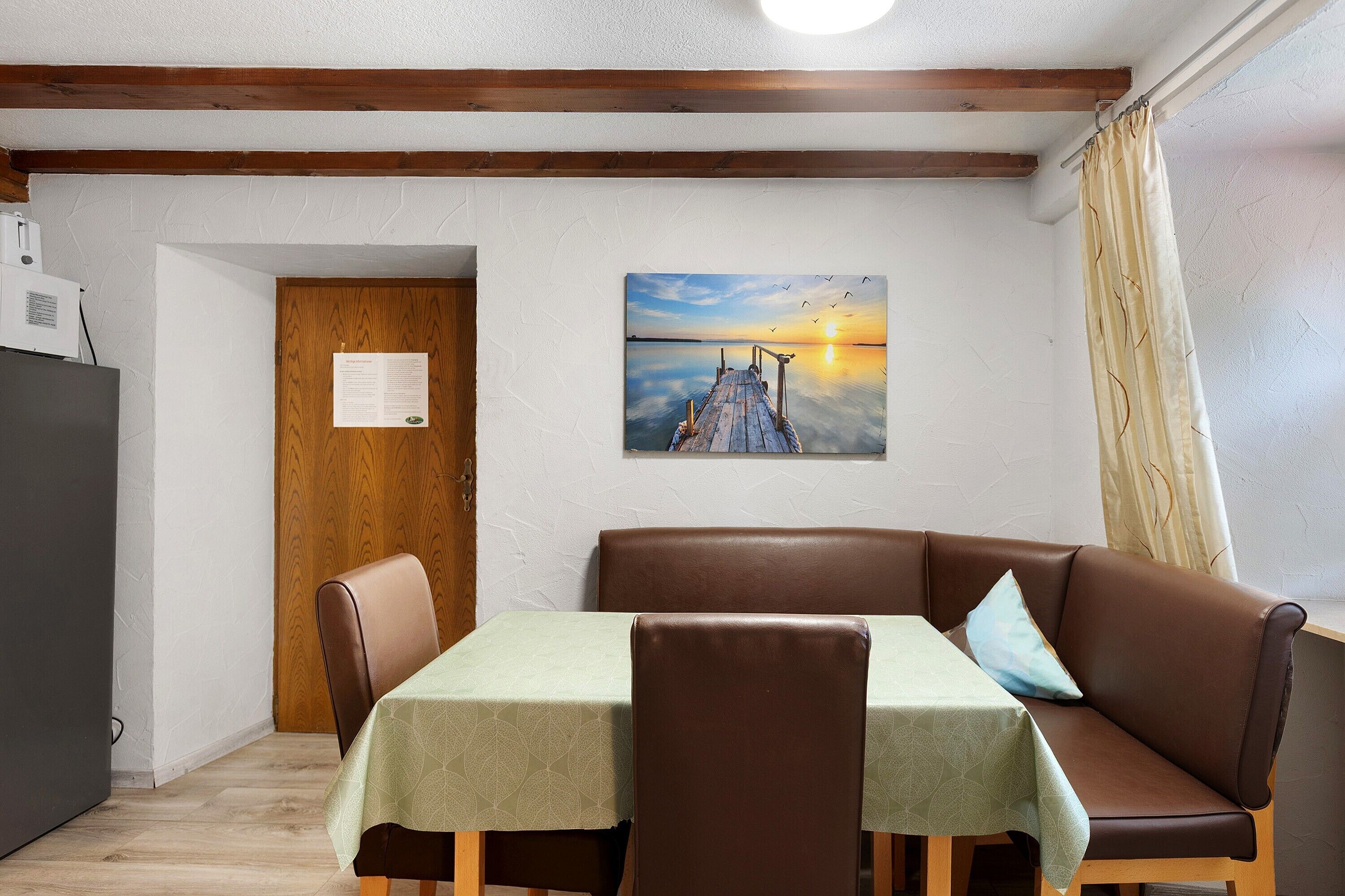 Ein Zimmer mit Holzboden, einem Tisch mit einer Tischdecke mit Blattmuster, einem braunen Ledersofa und einem Wandbild einer untergehenden Sonne.