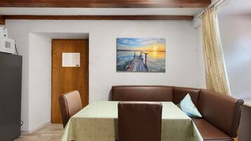 Ein Zimmer mit Holzboden, einem Tisch mit einer Tischdecke mit Blattmuster, einem braunen Ledersofa und einem Wandbild einer untergehenden Sonne.