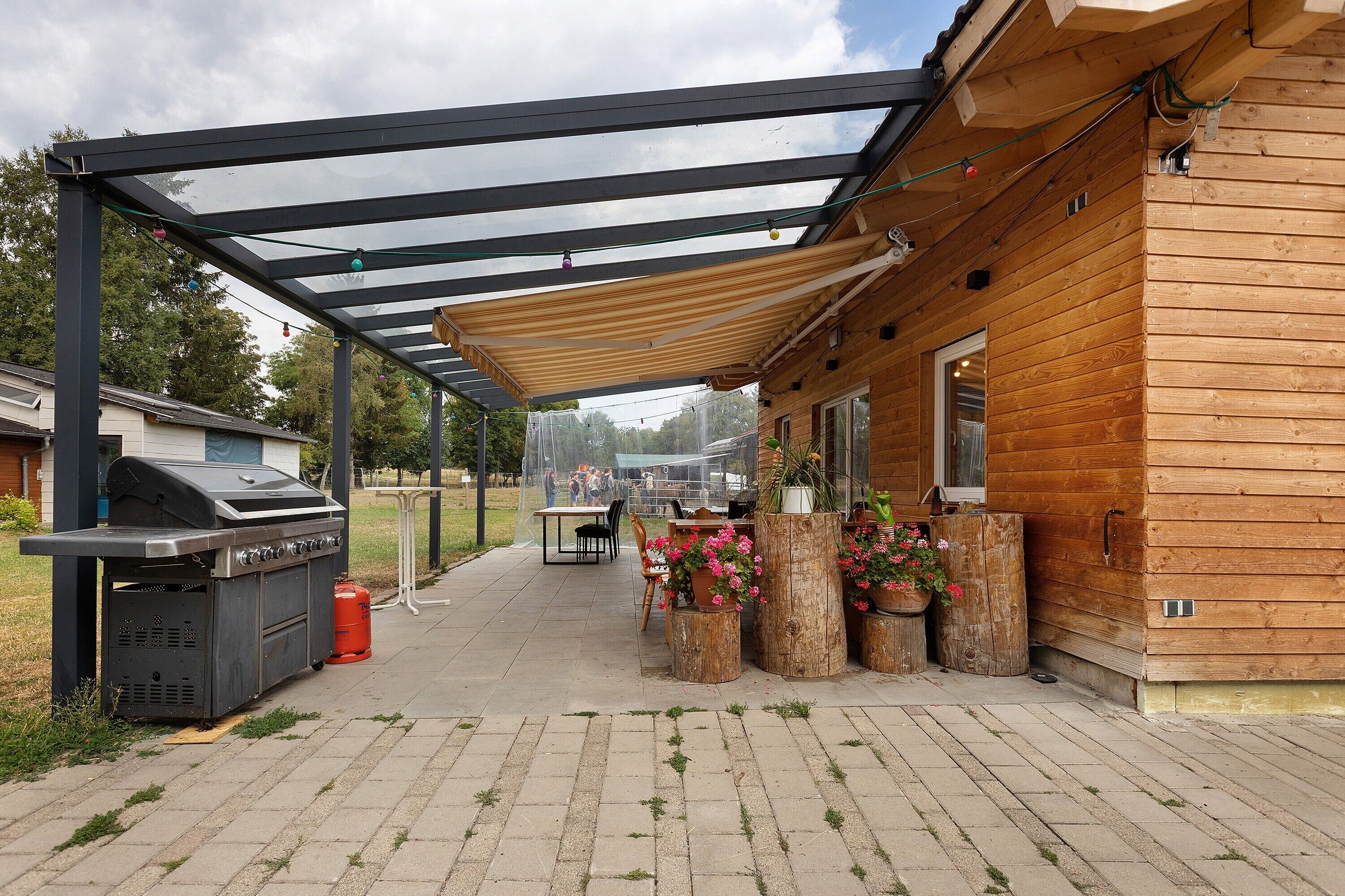 Eine überdachte Terrasse im Freien mit Grill, Blumentöpfen und einem Holzbau.