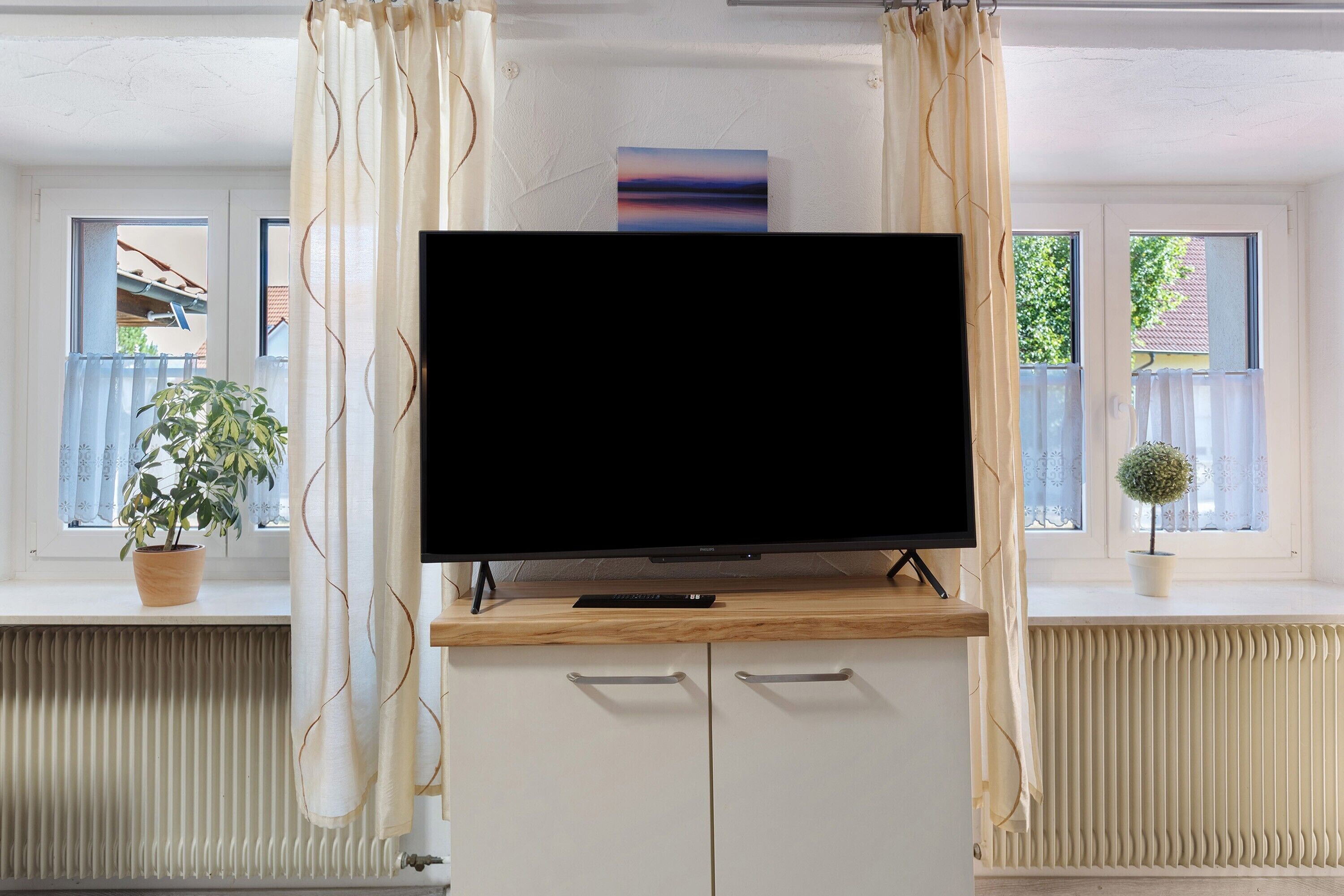Ein Fernseher auf einem Holzboden in einem Raum mit weißen Wänden, einer Heizung und zwei mit Vorhängen versehenen Fenstern.