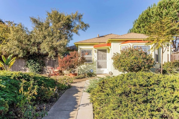 A residential house with a front porch, a pathway leading to the entrance, and a variety of plants and trees surrounding the property. - Wine Country Bungalow 2Bd/1Ba (Lompoc)