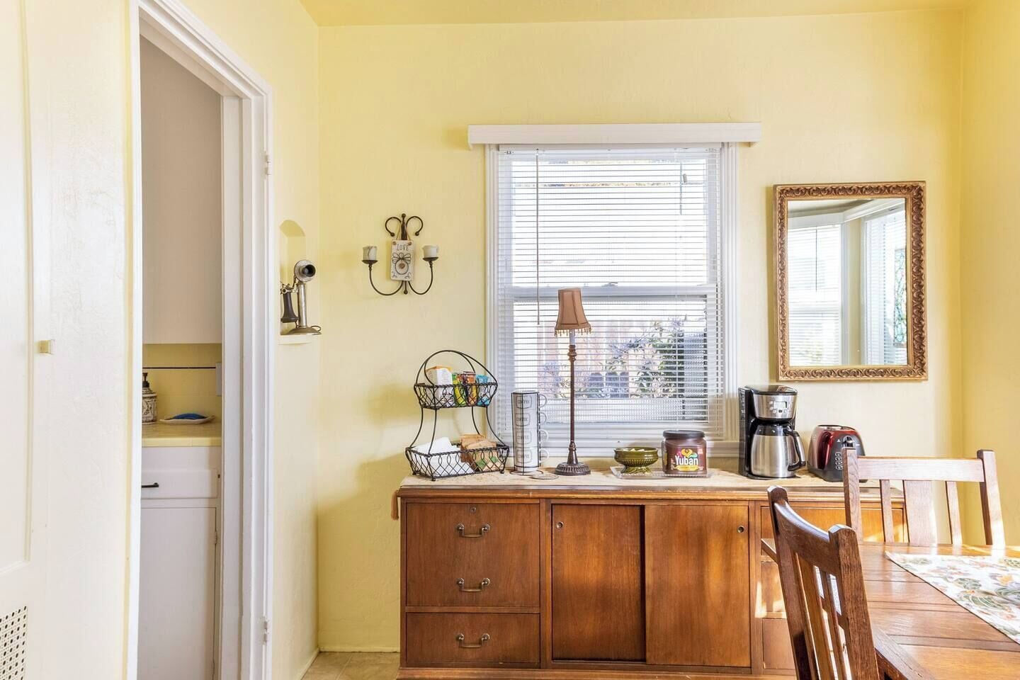 Un coin repas avec une table en bois, des chaises, une fenêtre avec des stores, une lampe, une machine à café et un miroir au mur.