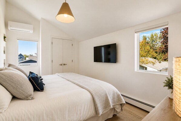A bedroom with a bed, a television, a window with a view, and an air conditioning unit.
