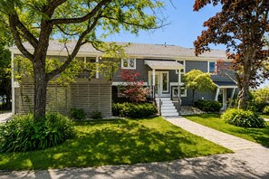 A modern two-story house with a well-maintained lawn, a pathway leading to the front door, and a variety of shrubs and trees.