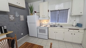A compact kitchen with white appliances, a window with blinds, and a small dining area with a wooden table and chair.