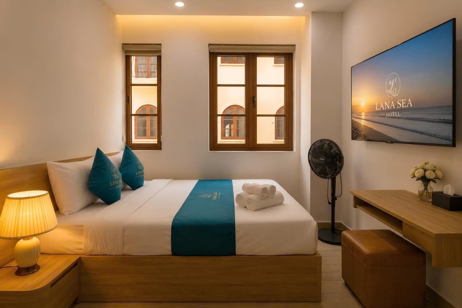 A hotel room with a bed, a desk, a chair, a standing fan, and a TV displaying a beach scene.