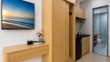 A modern hotel room with a TV displaying a scenic view, a wooden wardrobe, a kitchen area, and a glass shower enclosure.