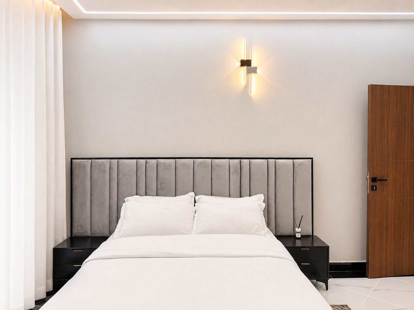 A modern bedroom with a bed, nightstands, a wall-mounted light fixture, and a wooden door.