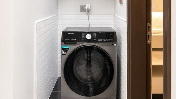 A compact, modern laundry room with a front-loading washing machine, a wall-mounted shelf above, and a door leading to another room.