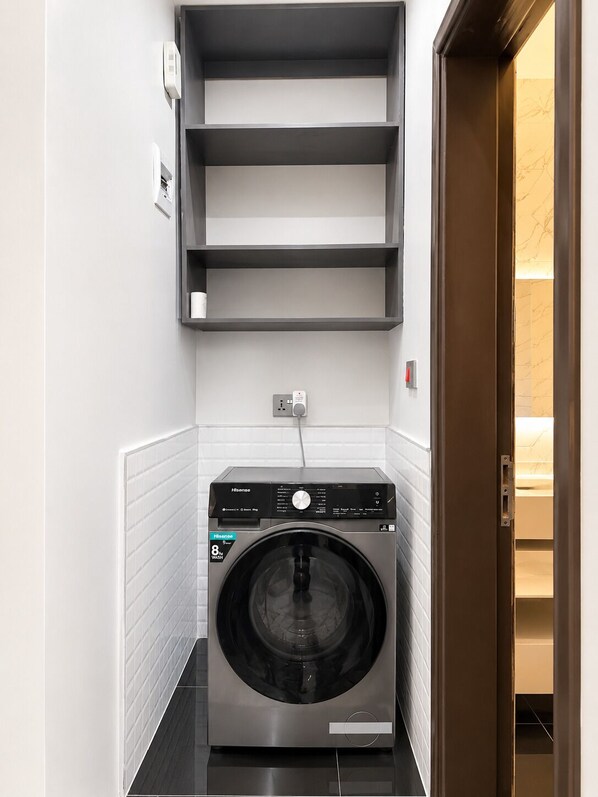 A compact, modern laundry room with a front-loading washing machine, a wall-mounted shelf above, and a door leading to another room.