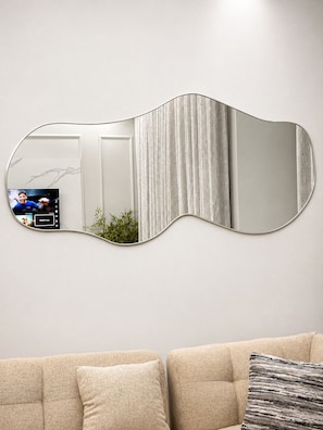 A modern living room with a unique curved mirror, a television displaying a live stream, and a sofa with cushions.