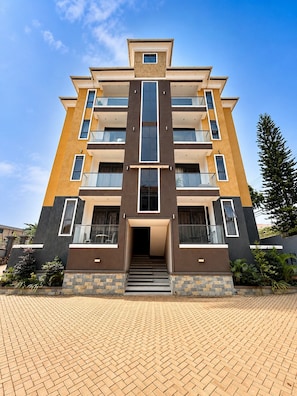 A multi-story building with a central entrance, balconies, and a brick-paved ground.