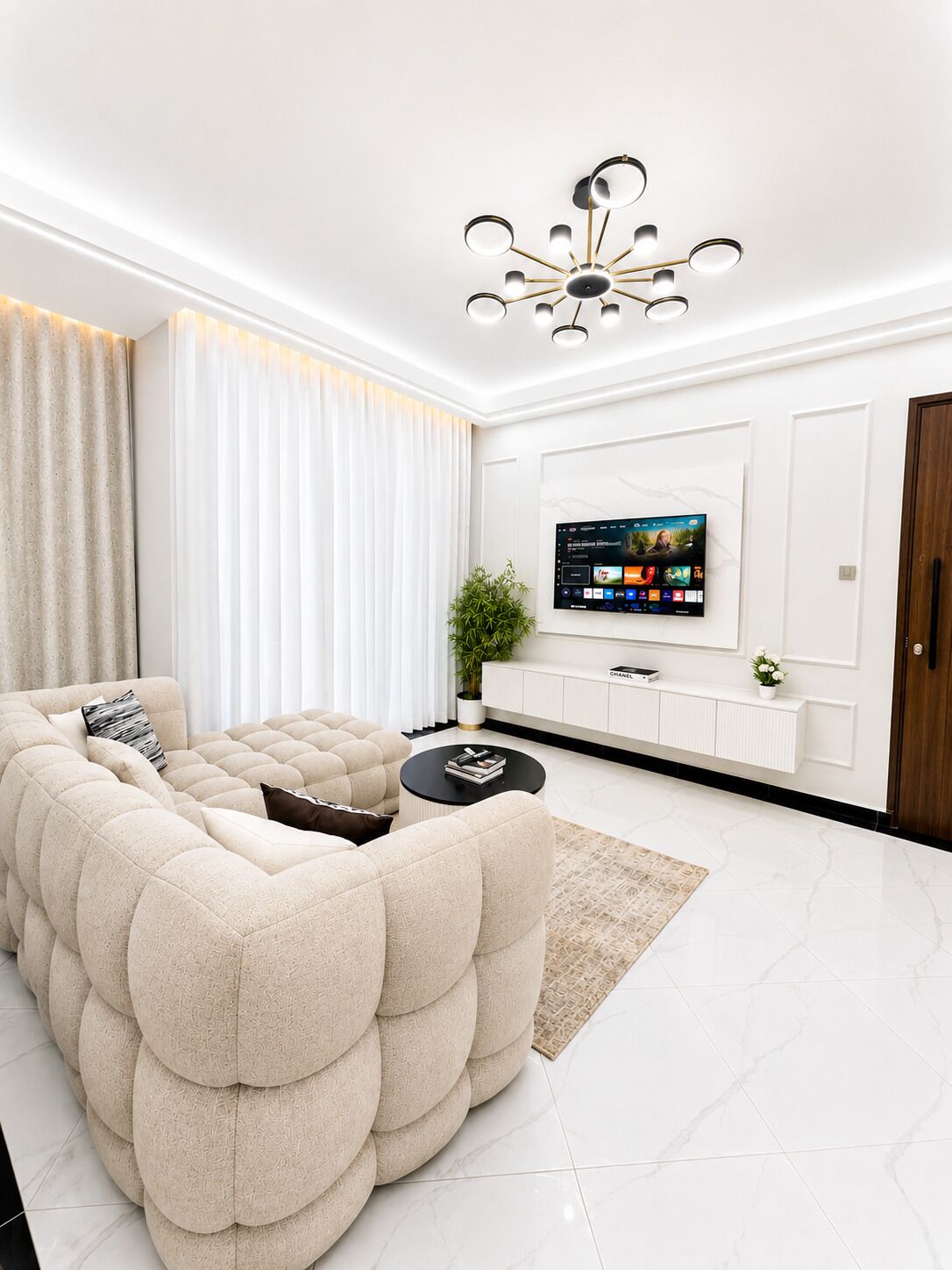 Un salon moderne avec un canapé d’angle, un téléviseur à écran plat fixé au mur, une table basse noire et un luminaire décoratif au plafond.