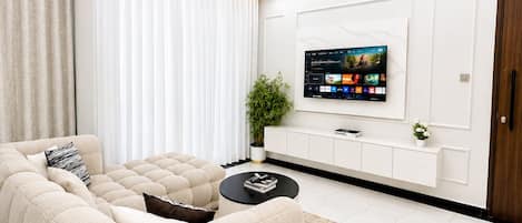 A modern living room with a sectional sofa, a flat-screen TV mounted on the wall, a black coffee table, and a decorative ceiling light.