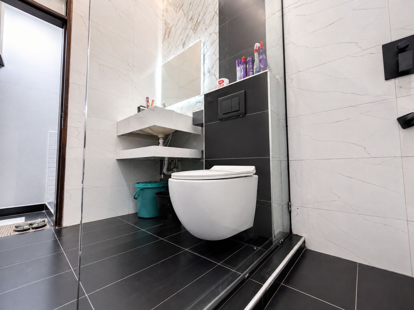 Un baño moderno con inodoro blanco, baldosas negras, un bote de basura verde agua y un lavamanos negro.