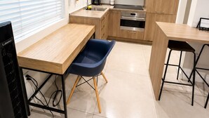 Een moderne keuken met witte kastjes, een roestvrijstalen spoelbak en een ingebouwde oven. Er staat een houten tafel met een blauwe stoel, en een zwart beeldscherm op een standaard.