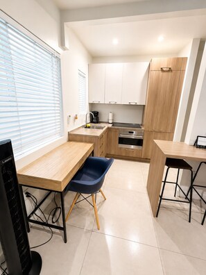 A modern kitchen with white cabinets, a stainless steel sink, and a built-in oven. There is a wooden table with a blue chair, and a black monitor on a stand. - Beautiful studio with 2 beds- Midtown (Miami)