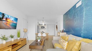 A modern living room with a flat-screen TV, a sofa with patterned cushions, a wooden coffee table, and a dining area with a wooden table and chairs. - Cozy 1BR- Bay View - Resort Pool & Private Balcony (Hallandale Beach)