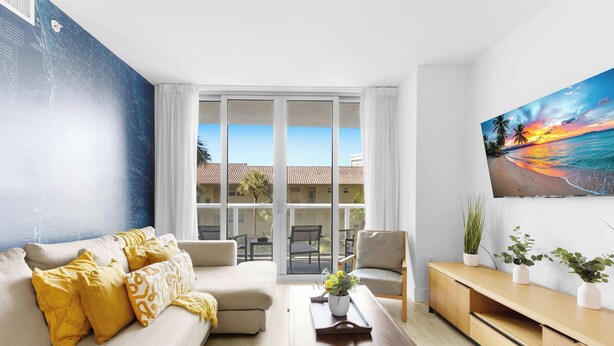 A modern living room with a sofa, a coffee table, and a painting of a beach scene. - Cozy 1BR- Bay View - Resort Pool & Private Balcony (Hallandale Beach)