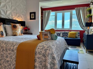 A well-lit bedroom with a bed, a bedside table, a nightstand, a window with curtains, a sofa, and a small table.