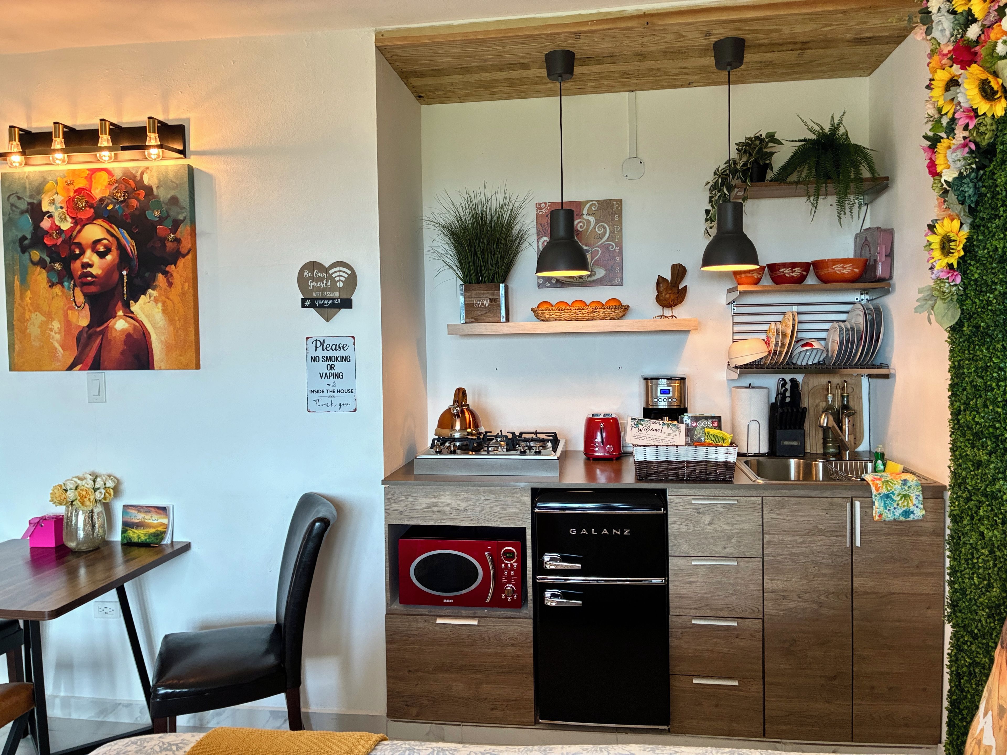 Une cuisine moderne équipée d’un four à micro-ondes, d’une cuisinière et d’un lave-vaisselle. On y trouve des luminaires suspendus, une plante et une étagère murale où sont rangés de la vaisselle.