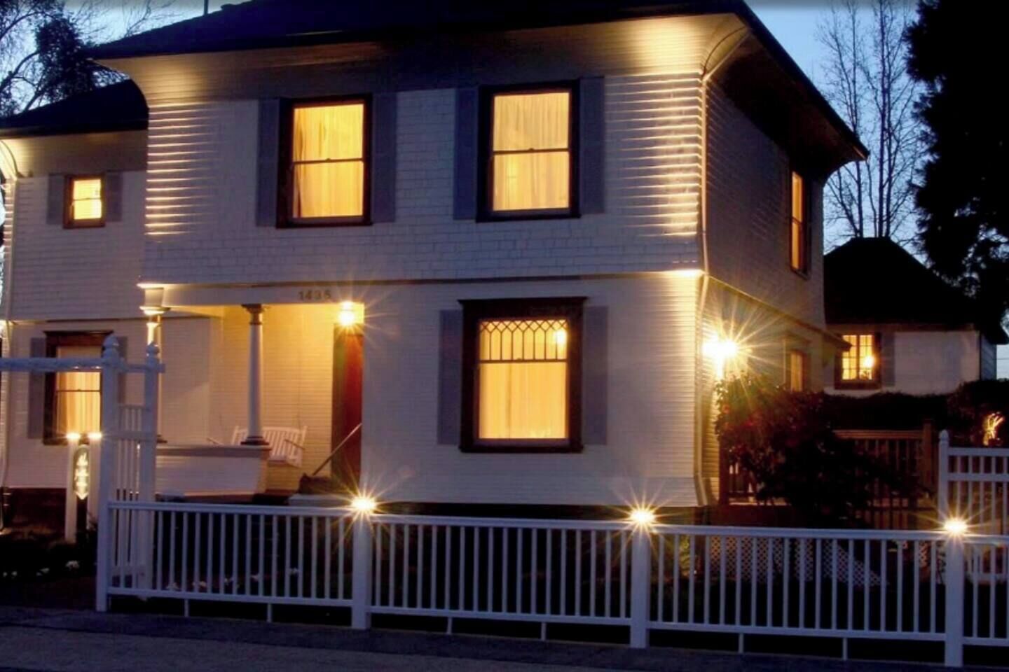 Een twee verdiepingen tellend huis met een witte gevel, een voortuin met een witte balustrade en meerdere ramen met licht.