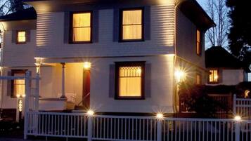 Een twee verdiepingen tellend huis met een witte gevel, een voortuin met een witte balustrade en meerdere ramen met licht.