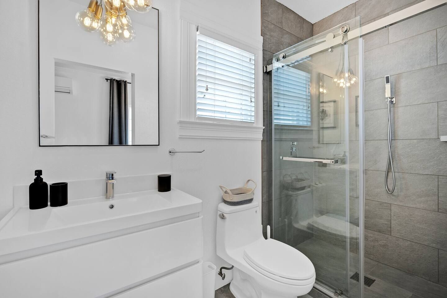 A modern bathroom with a white vanity, a toilet, a glass-enclosed shower, and a chandelier.