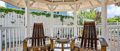 A covered outdoor seating area with two wooden chairs and a small table, surrounded by white railings and greenery.